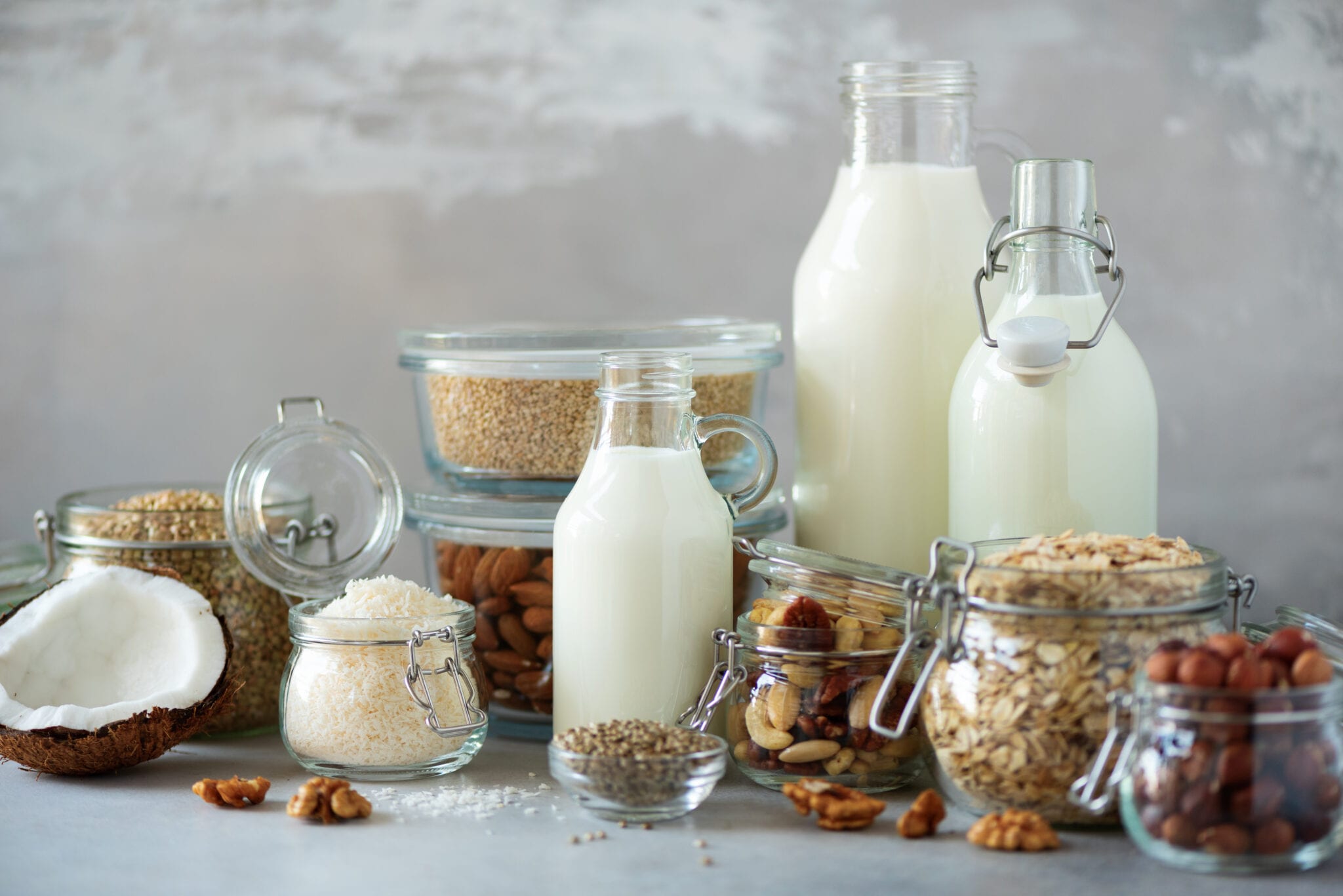 Glass Bottles Of Vegan Plant Milk And Almonds, Nuts, Coconut, Hemp Seed Milk On Grey Concrete Background