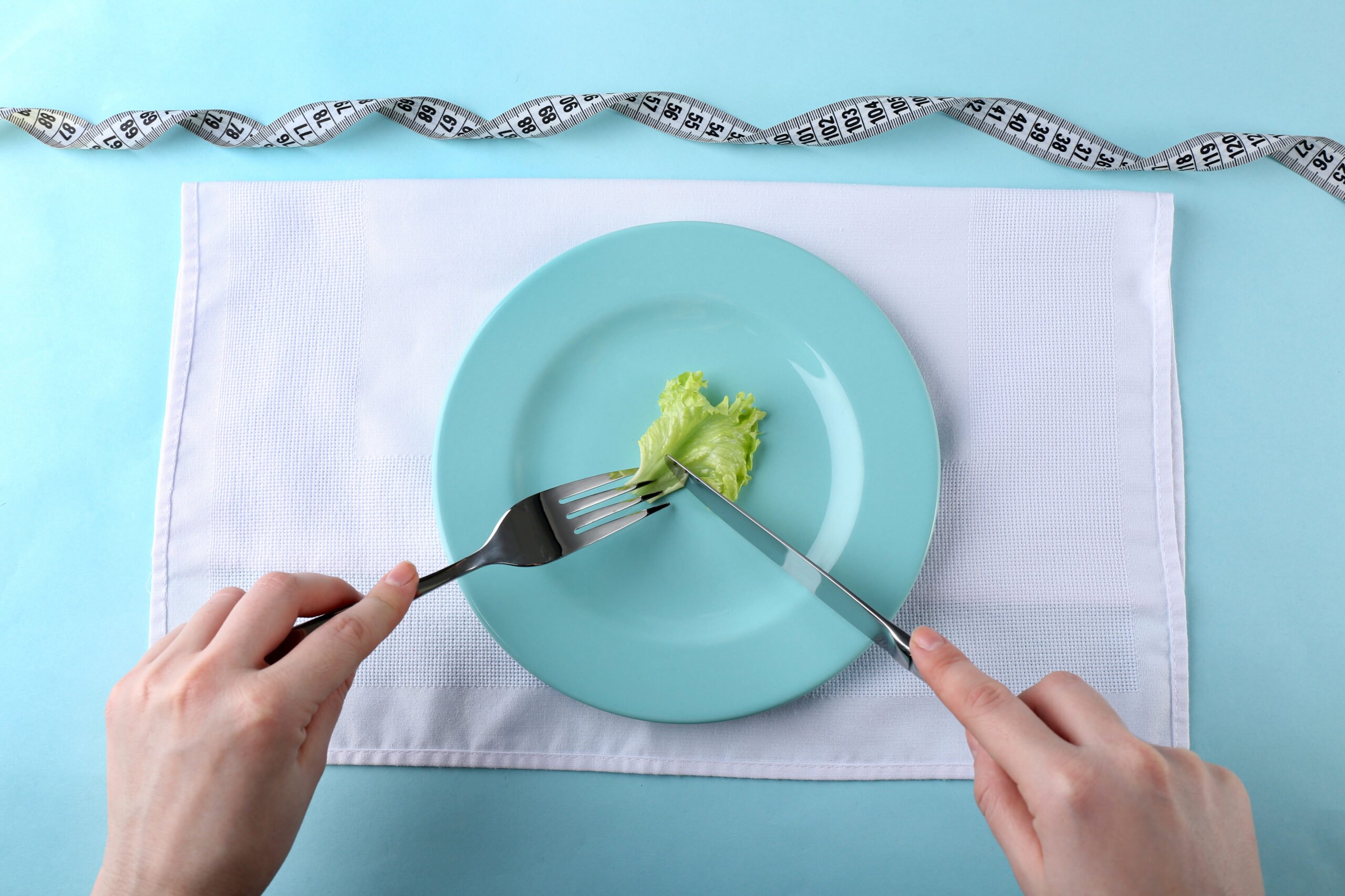 Picture of female hands holding knife and fork on plate cutting piece of lettuce leaf, top view