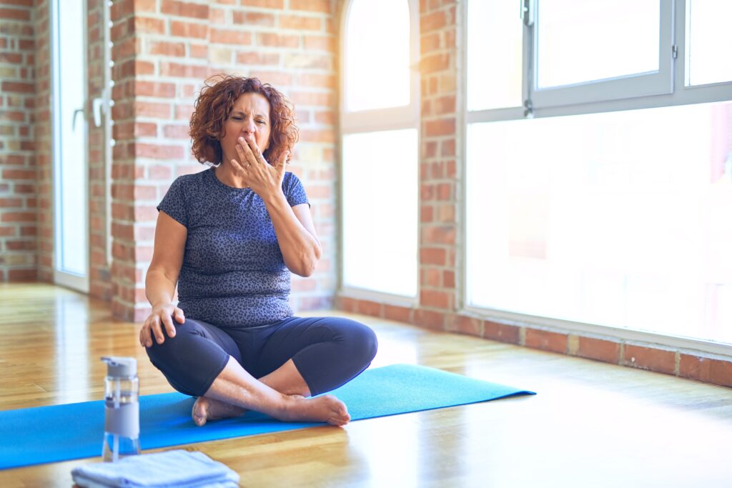 middle age beautiful sportswoman wearing sportswear sitting on mat practicing yoga at home bored yawning tired covering mouth with hand. restless and sleepiness.