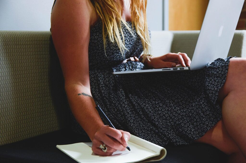 woman sitting on couch working writing in notebook and on laptop